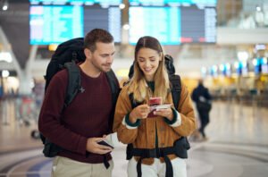 Happy couple, airport and phone with ticket, travel app and adventure with excited face, conversation and smile. Man, woman and smartphone for digital booking of hotel, taxi or bus for transportation